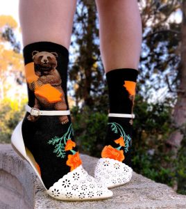 These black crew socks by ModSocks have a bear hugging the orange state of California with a big smile on his face. On the foot the California state flower - the California poppy - blooms beautifully. The look is styles with white flats with person standing on concrete wall.