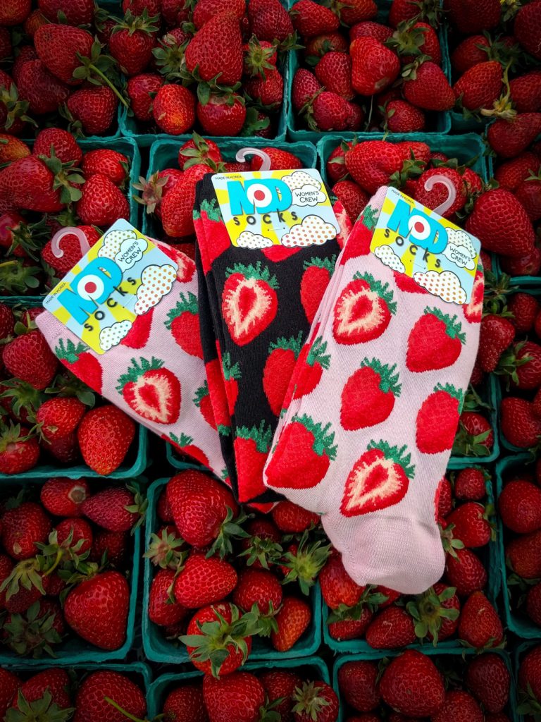 Strawberry crew socks in petal pink and black designed by ModSocks sit on top of strawberry baskets at the summertime farmer's market in the pacific northwest.
