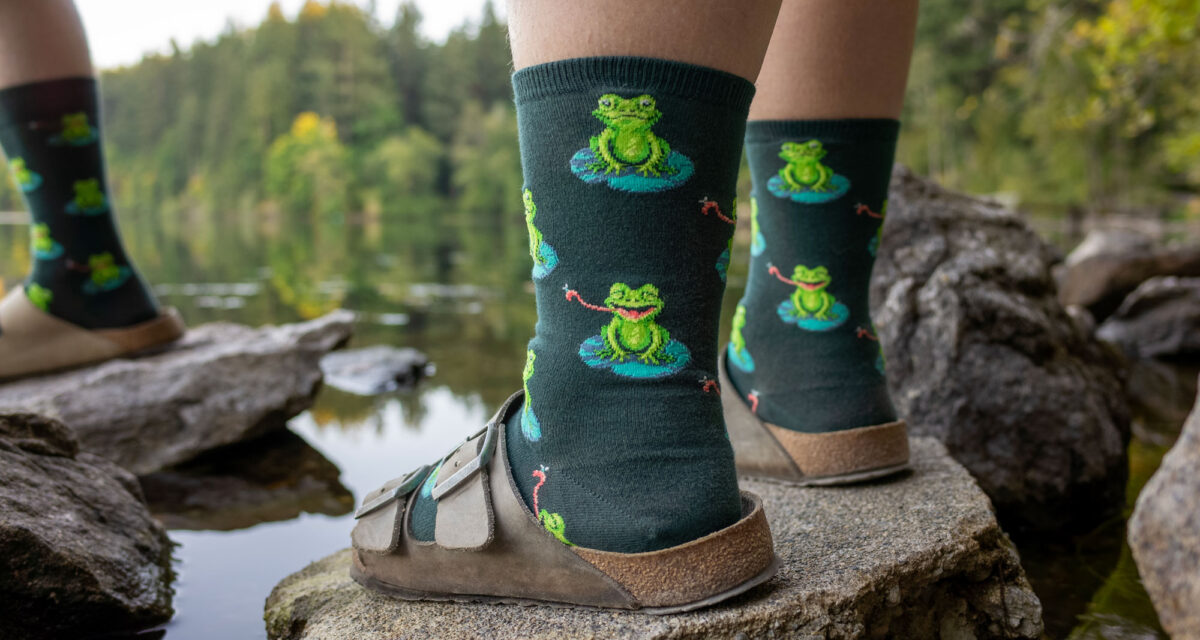 A model wears frog socks with sandals and stands on a rock beside a lake surrounded by trees.